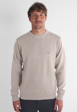 Pull en tricot beige avec col rond, poignets et ourlet côtelés. Petit emblème bleu sur la poitrine. Texture douce et coupe régulière.
