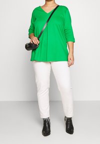 Grünes V-Ausschnitt Langarmshirt mit lockerer Passform, kombiniert mit cremefarbenen, tapered Pants und schwarzen Knöchelstiefeln mit spitzem Absatz.