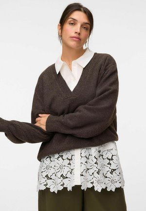 Donna che indossa una camicia bianca con collo e orlo in pizzo floreale, un maglione scuro a V e pantaloni verde oliva su sfondo semplice.