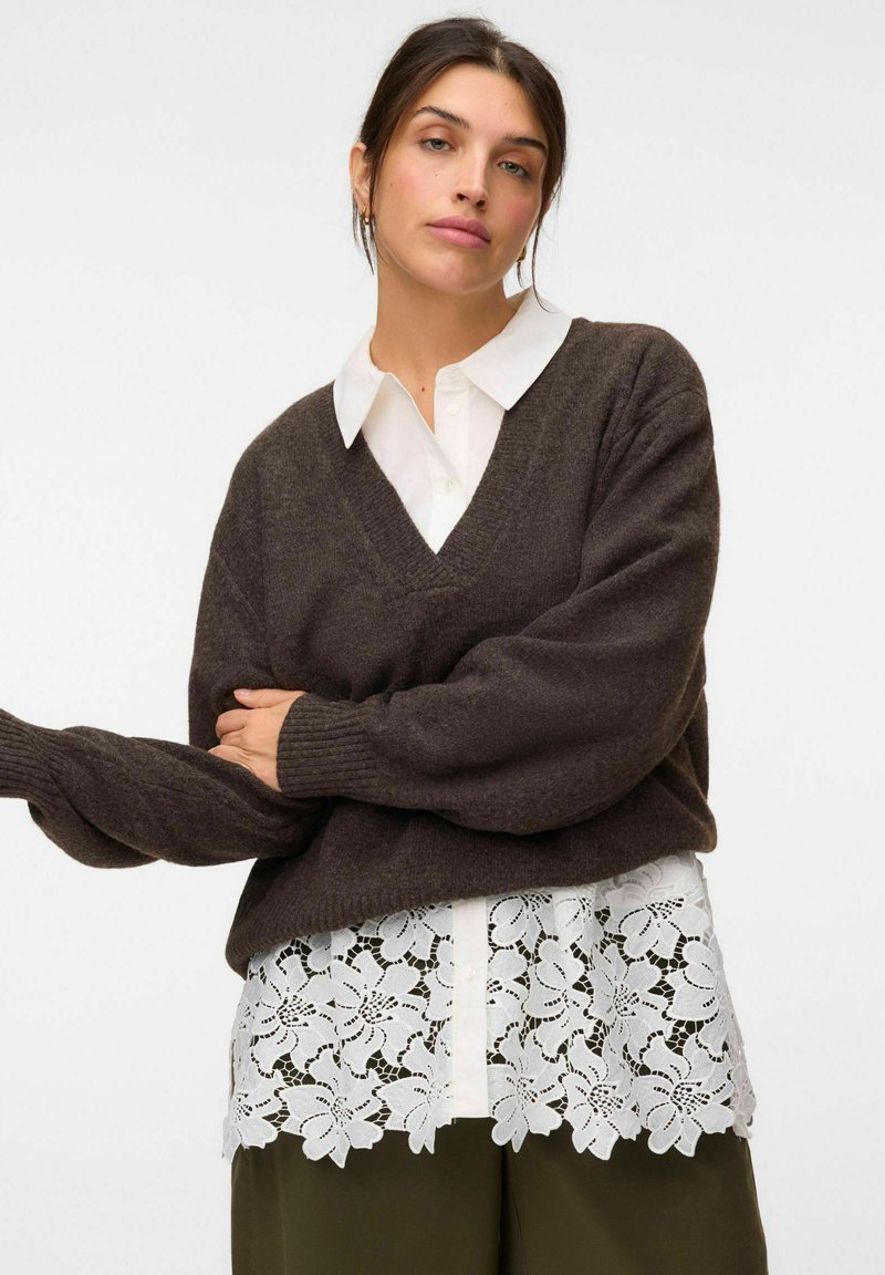 Femme portant une chemise blanche à col avec un ourlet en dentelle florale, un pull col V marron foncé et un pantalon vert olive sur un fond uni.