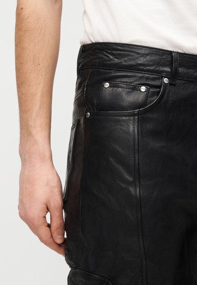 Ανδρικό χέρι που στηρίζεται δίπλα σε μαύρο δερμάτινο παντελόνι με ορατή μπροστινή τσέπη και λευκό πουκάμισο που έχει μπει μέσα.