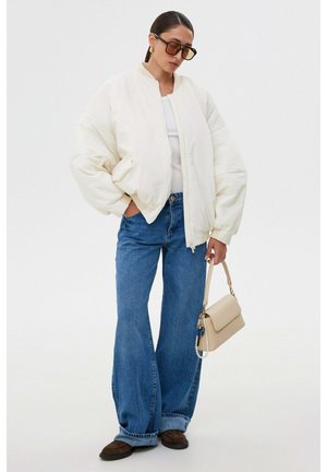 Kobieta w białej, dużej kurtce puchowej, białym topie, szerokich niebieskich jeansach, brązowych butach i okularach przeciwsłonecznych, trzymająca beżową torebkę z perłową ozdobą.