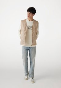 Beige gesteppte Weste über einem cremefarbenen Sweatshirt, hellblauen Jeans und weißen Sneakers. Die Weste hat einen Reißverschluss und seitliche Taschen.