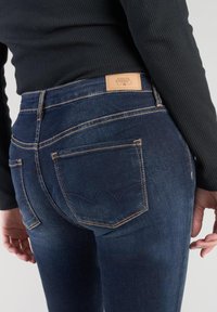 Jeans de mezclilla azul oscuro con cintura alta y un ligero desvanecimiento. Cuentan con dos bolsillos traseros con costura decorativa y una etiqueta de cuero marrón.