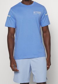 Camiseta deportiva de color azul claro con mangas cortas, que presenta un pequeño logo blanco y sutiles detalles en los hombros. Tejido suave con un ajuste relajado.