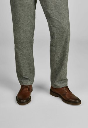 Braune Lederschuhe mit Schnürdetails, kombiniert mit grauen strukturierten Hosen. Die glatte, glänzende Oberfläche der Schuhe steht im Kontrast zum Gewebe der Hose.