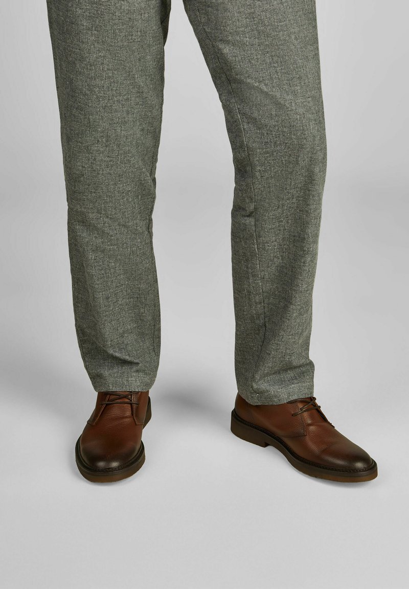 Bruine leren schoenen met vetersluiting, gecombineerd met grijze gestructureerde broek. Het gladde, glanzende oppervlak van de schoenen contrasteert met de weving van de stof.