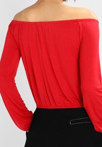 Rode off-the-shoulder top met lange mouwen, geplooide tailleband en een zachte textuur. De achterkant heeft een relaxte pasvorm met een elastische zoom.