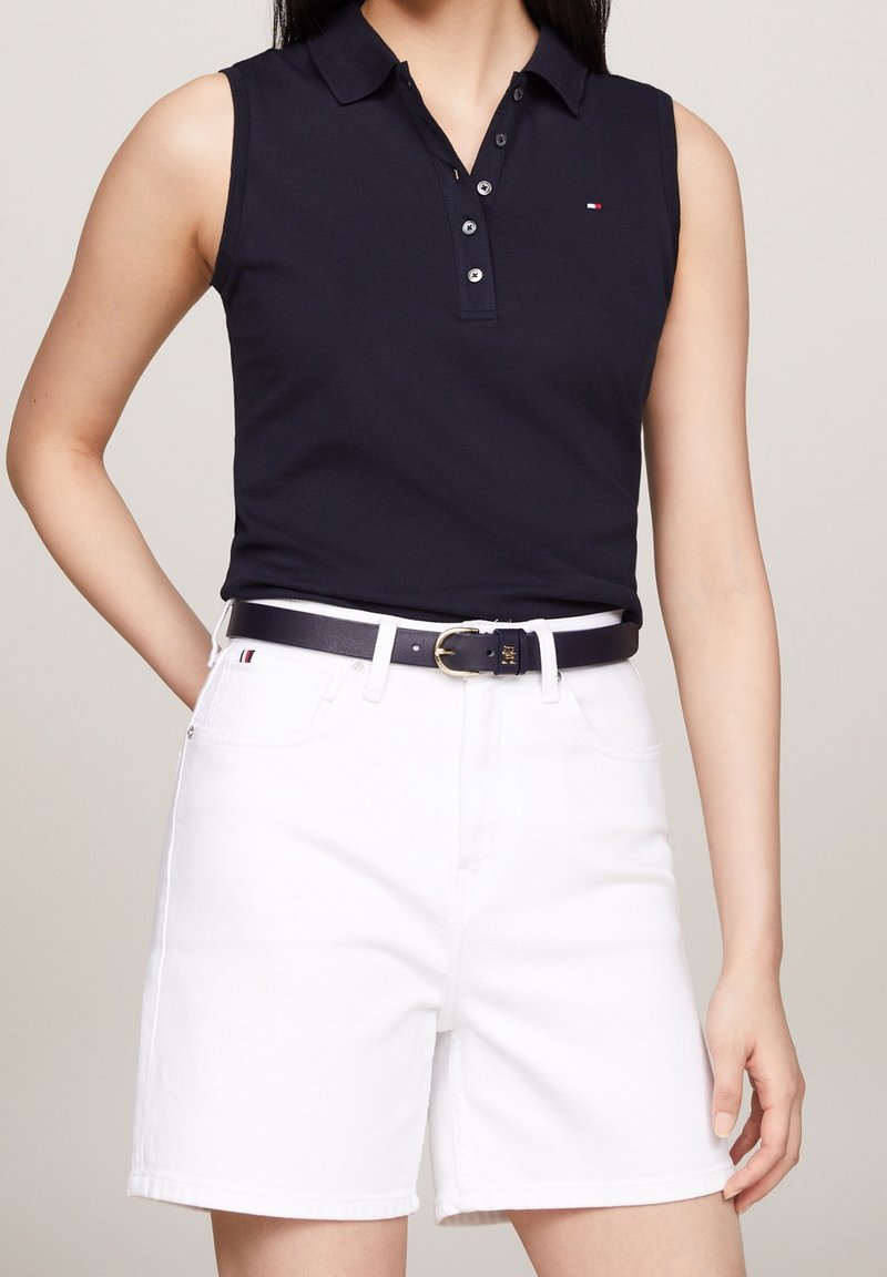 Personne portant un polo noir sans manches et un short blanc avec une ceinture noire, montrée des épaules jusqu'au milieu des cuisses sur fond uni.