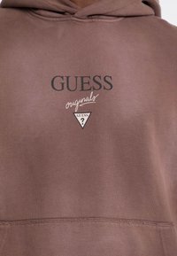 Felpa marrone con tasca a canguro, logo nero "GUESS", "originals" in corsivo e una patch triangolare con un punto interrogativo.