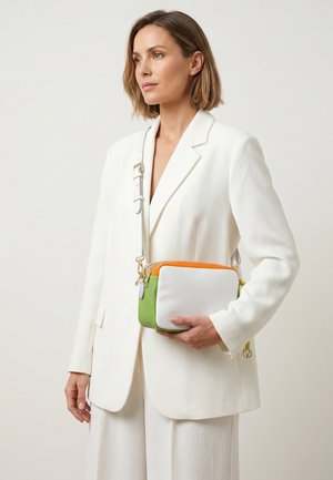 Vrouw in wit pak houdt een witte, groene en oranje schoudertas met een gouden ritssluiting en verstelbare band tegen een neutrale achtergrond.