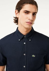 Camicia azzurra a maniche corte con bottoni in cotone, dotata di una tasca anteriore e di un piccolo logo di coccodrillo verde sul lato sinistro del petto. Texture liscia.