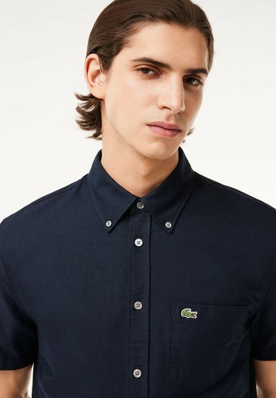 Chemise à manches courtes bleu marine, fabriquée en coton, avec une poche frontale et un petit logo crocodile vert sur la poitrine gauche. Texture lisse.