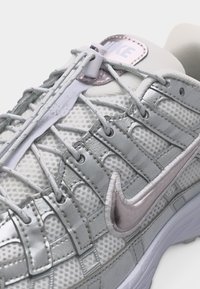 Κοντινό πλάνο ενός ασημί και λευκού sneaker Nike με ύφασμα από πλέγμα, συνθετικές επιστρώσεις και σύστημα κλεισίματος κορδονιών με ταχυσύνδεσμο σε λευκή σόλα.