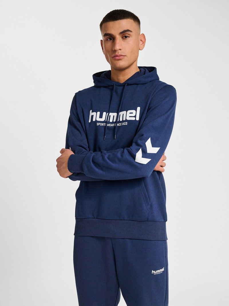 Jonge man staat met armen over elkaar, draagt een marineblauwe Hummel hoodie en bijpassende joggingbroek met witte logotekst en chevrons op de mouw.