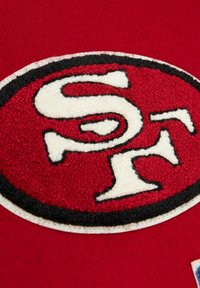 Roter Stoff mit einem runden Aufnäher, der mit einem weißen "SF"-Logo umrandet in Schwarz gestaltet ist. Die Textur ist filzartig und das Design ist kräftig und klar.