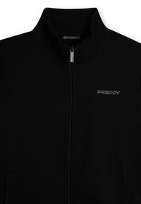 Chaqueta negra con cremallera y cuello alto, hecha de tela suave. Cuenta con bolsillos laterales y un sutil logo gris de Freddy en el pecho izquierdo.