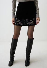 Mini gonna in velluto nero con ricami floreali all'orlo, abbinata a stivali in pelle neri fino al ginocchio e collant neri trasparenti.