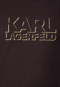 Czarna tkanina z wypukłym, wyciętym na złoto logo "KARL LAGERFELD". Gładka faktura, wyraźna czcionka, centralne umiejscowienie, minimalistyczny design.
