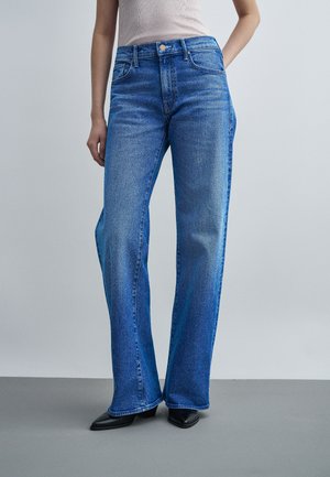 Žena má na sebe modré nohavice s vysokým pásom a širokými nohavicami a svetlo béžové tielko bez rukávov, stojí s jednou rukou na boku na jednofarebnom pozadí.