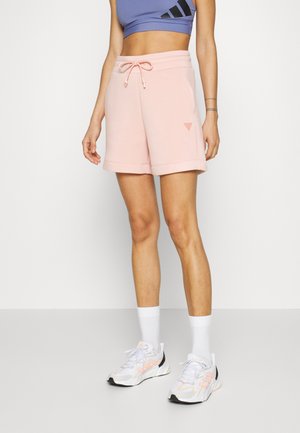 Femme portant un short à cordon rose clair, des chaussures de sport blanches, des chaussettes blanches et un haut de sport court violet avec un logo noir.