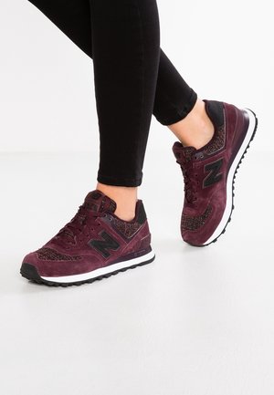 Personne portant un pantalon noir et des baskets New Balance bordeaux marchant sur un sol blanc.