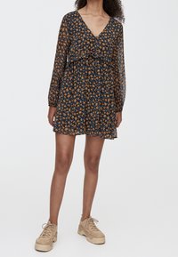 Robe bleu marine à motifs floraux avec un décolleté en V et des manches longues transparentes. Détail à volants à la taille, assortie à des baskets beiges.