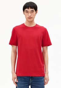 Junger Mann mit kurzem schwarzen Haar, der ein schlichtes rotes T-Shirt mit kurzen Ärmeln und blaue Jeans trägt, vor einem weißen Hintergrund.