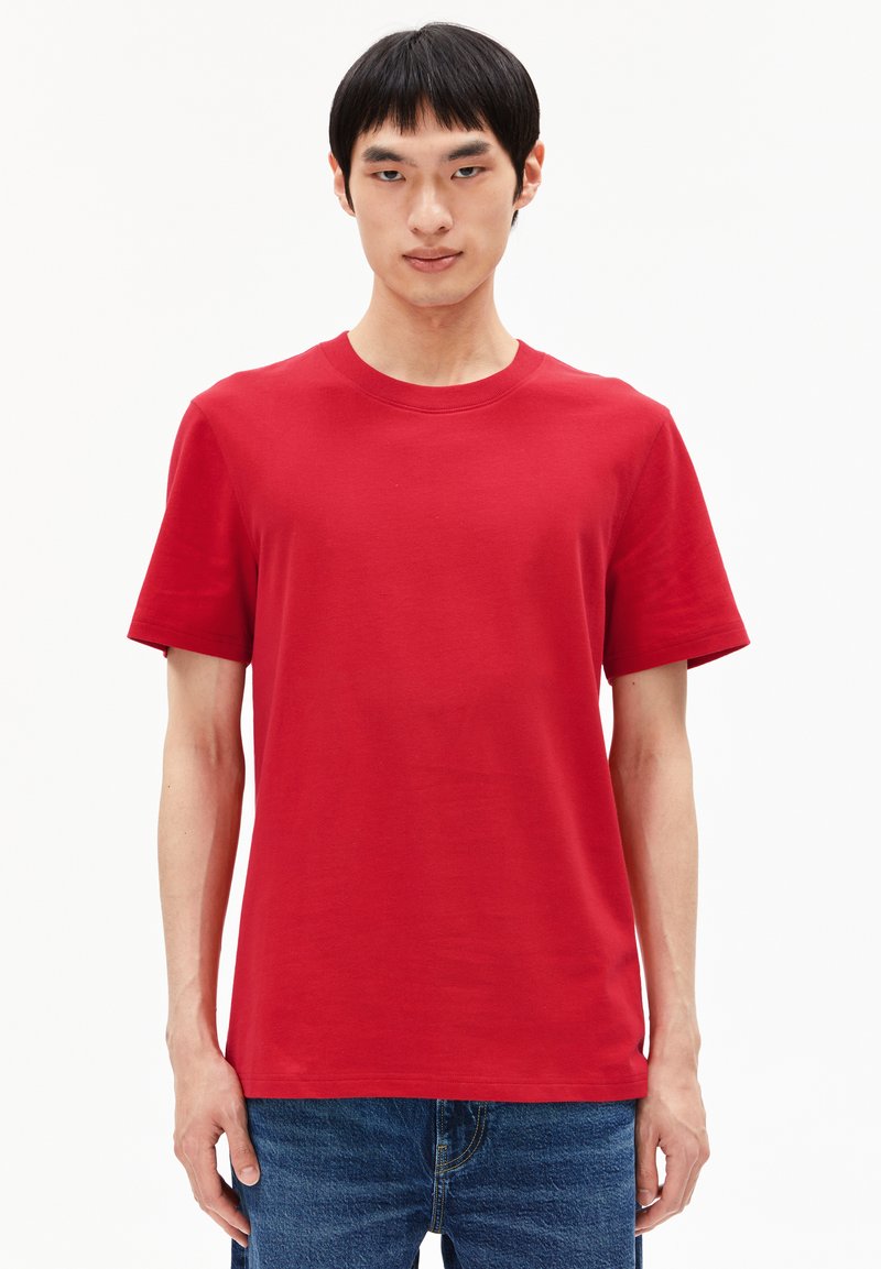 Junger Mann mit kurzem schwarzen Haar, der ein schlichtes rotes T-Shirt mit kurzen Ärmeln und blaue Jeans trägt, vor einem weißen Hintergrund.