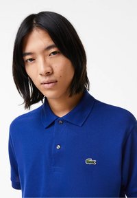 Jeune personne aux cheveux noirs mi-longs portant un polo bleu Lacoste avec un logo de crocodile sur fond blanc.