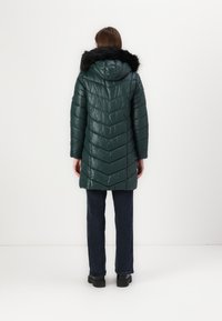 Cappotto puffy verde con cappuccio e rifiniture in pelliccia sintetica, caratterizzato da un motivo a chevron trapuntato e una texture lucida, abbinato a jeans scuri e stivali.