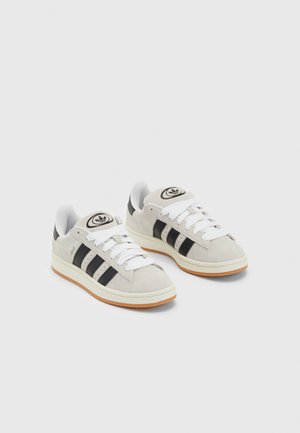 Adidași din suede gri cu șireți albi, dungi negre și talpă din cauciuc alb. Prezintă un logo circular pe limbă și o talpă din cauciuc.