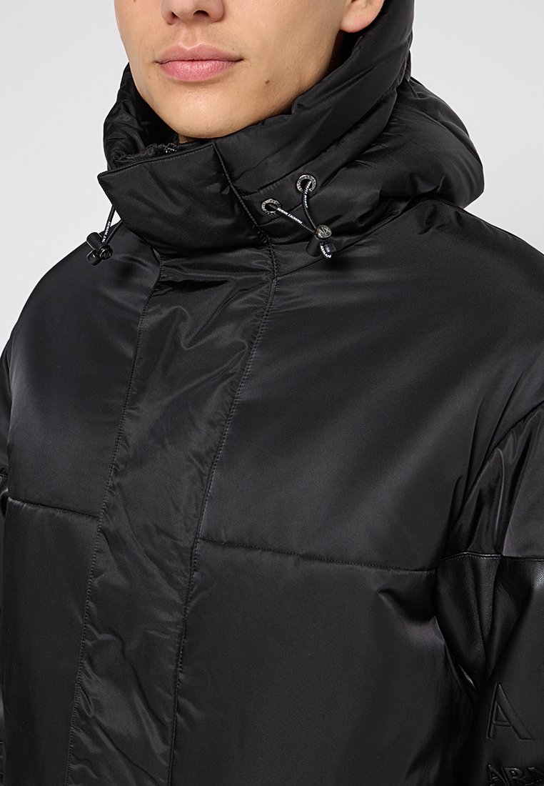 Giacca imbottita nera con colletto alto, dettagli con cordino e una texture liscia. Presenta maniche simili alla pelle e un design del corpo trapuntato.