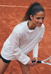 Langermet, hvit treningstrøye med rund hals, parret med svarte shorts. Bakgrunnen viser en tennisbane med oransje leire.