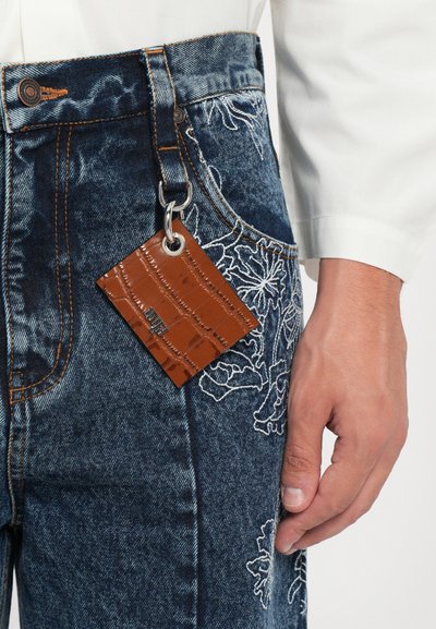 Etichetta in pelle marrone con motivo coccodrillo attaccata a jeans di denim con ricami floreali e cuciture arancioni su sfondo bianco.