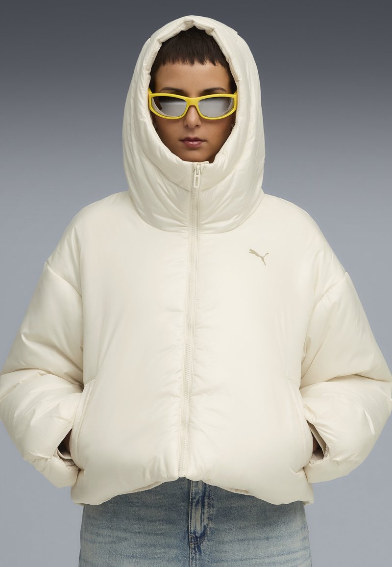 Puffjacka i off-white, med hög huva, dragkedjeslutning och ribbad mudd. Åtföljd av gula rektangulära solglasögon.