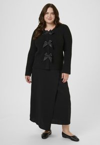 Schwarze Strickjacke mit drei dekorativen schwarzen Schleifen auf der Vorderseite, getragen über einem schwarzen Maxirock und schwarzen Schuhen.