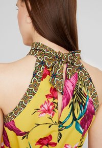 Floral halter top featuring a yellow base and vibrant pink and green patterns, with a bow tie closure and decorative collar detailing.