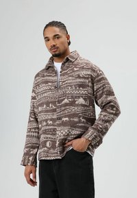 Bruine geruite flanellen overhemd met witte motieven; voorzien van een kraag, knoopsluiting en lange mouwen. Draagt zwart broek.