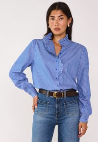 Blusa a righe blu e bianche con colletto arricciato e maniche lunghe, abbinata a jeans in denim a vita alta e una cintura marrone.