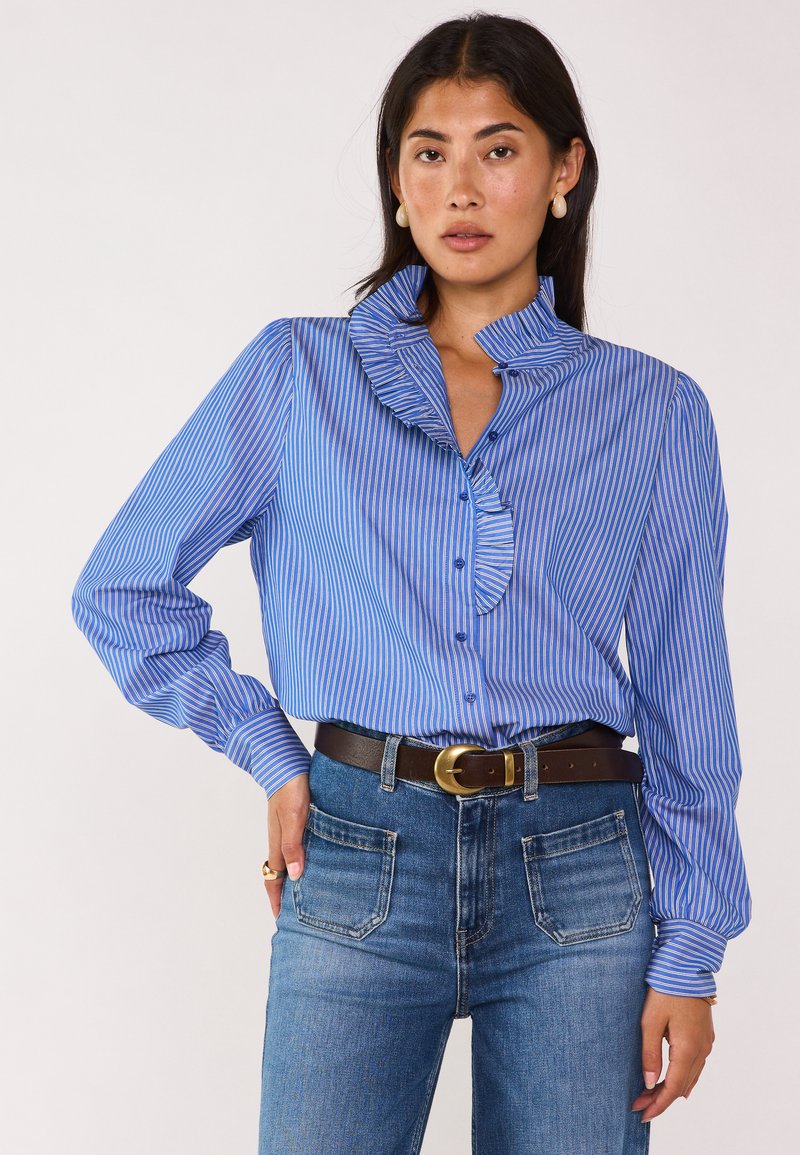 Blusa a righe blu e bianche con colletto arricciato e maniche lunghe, abbinata a jeans in denim a vita alta e una cintura marrone.