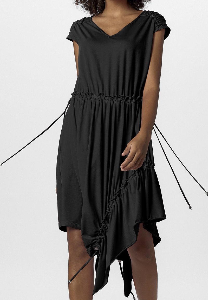 Femme portant une robe noire avec une taille froncée et un ourlet asymétrique, avec des cordons réglables aux épaules, à la taille et à la jupe.