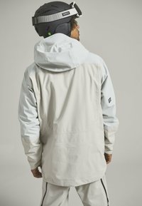 Lightweight, two-tone ski jacket in pale gray and light blue, with a durable texture, adjustable hood, and black logo accent on the sleeve.