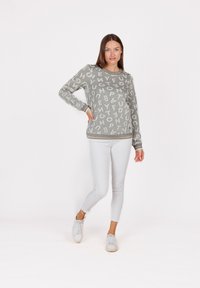 Donna che indossa un maglione grigio con lettere bianche, pantaloni bianchi aderenti e sneakers bianche, in piedi con una mano sulla vita.