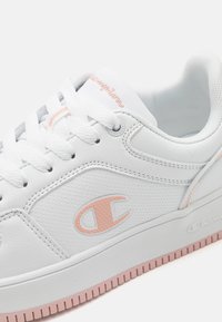 Zapatilla deportiva de cuero blanca con acentos en rosa. Presenta un panel lateral texturizado, logo en el lado exterior y suela de goma texturizada.