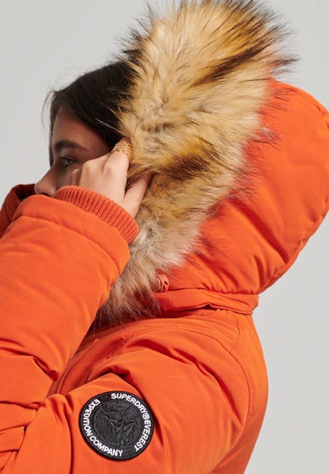 Oranje donsjack met een met bont gevoerde capuchon. Heeft een zwarte ronde patch op de linkermouw en ribgebreide manchetten. Glad, geïsoleerd materiaal.