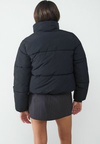 Czarna kurtka puchowa z wysokim kołnierzem, pikowanym wzorem i widocznym poziomym przeszyciem. Gładka tekstura i nieco luźniejszy krój.
