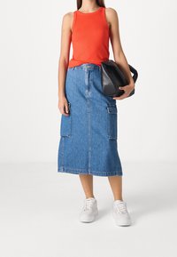 Top sin mangas rojo con textura acanalada, falda midi de denim con dos grandes bolsillos laterales, y zapatillas deportivas blancas de suela gruesa. Sosteniendo un bolso negro, de pie sobre un fondo blanco.