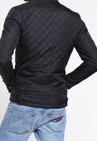Sort quiltet jakke med et diamantmønster, udstyret med stående krave, tætsiddende design og sidelommer, båret over blå jeans.