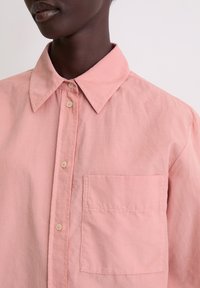 Light pink button-up shirt made of cotton, featuring a classic collar, front pocket, and subtle texture.